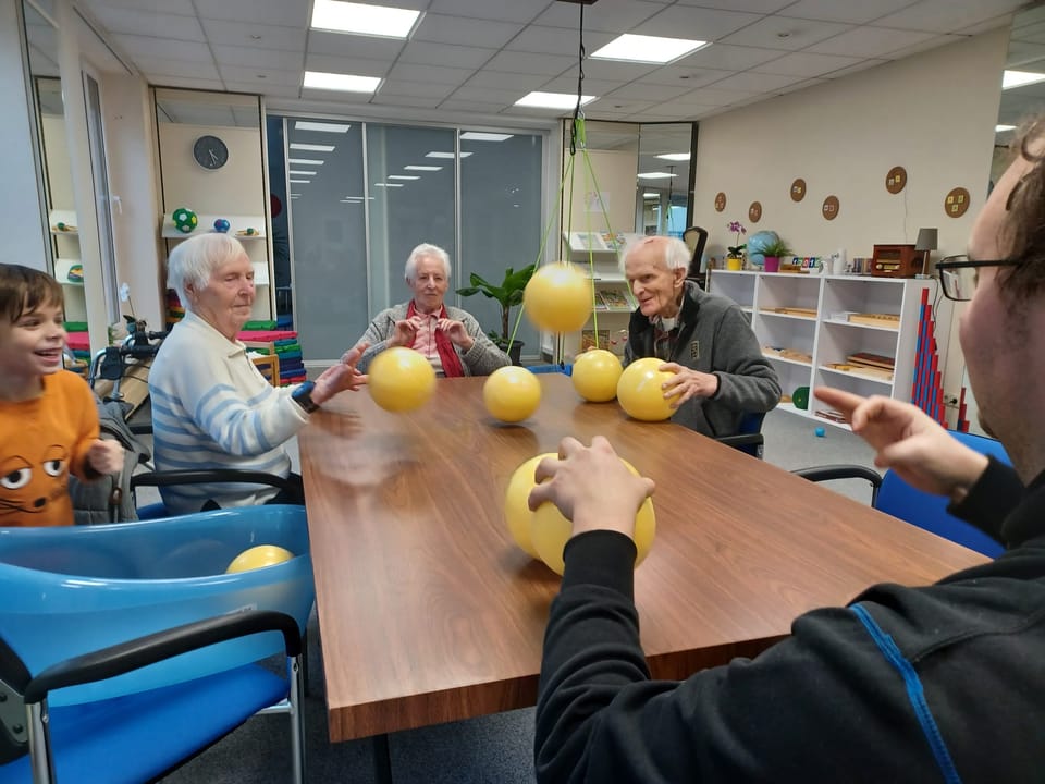 Senioren und ein Kind spielen sich am großen Tisch im Gruppenraum gelbe Bälle zu